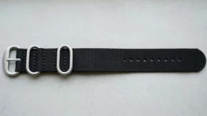 Black 3-ring Zulu strap with brushed steel oval hardware laid flat on neutral surface