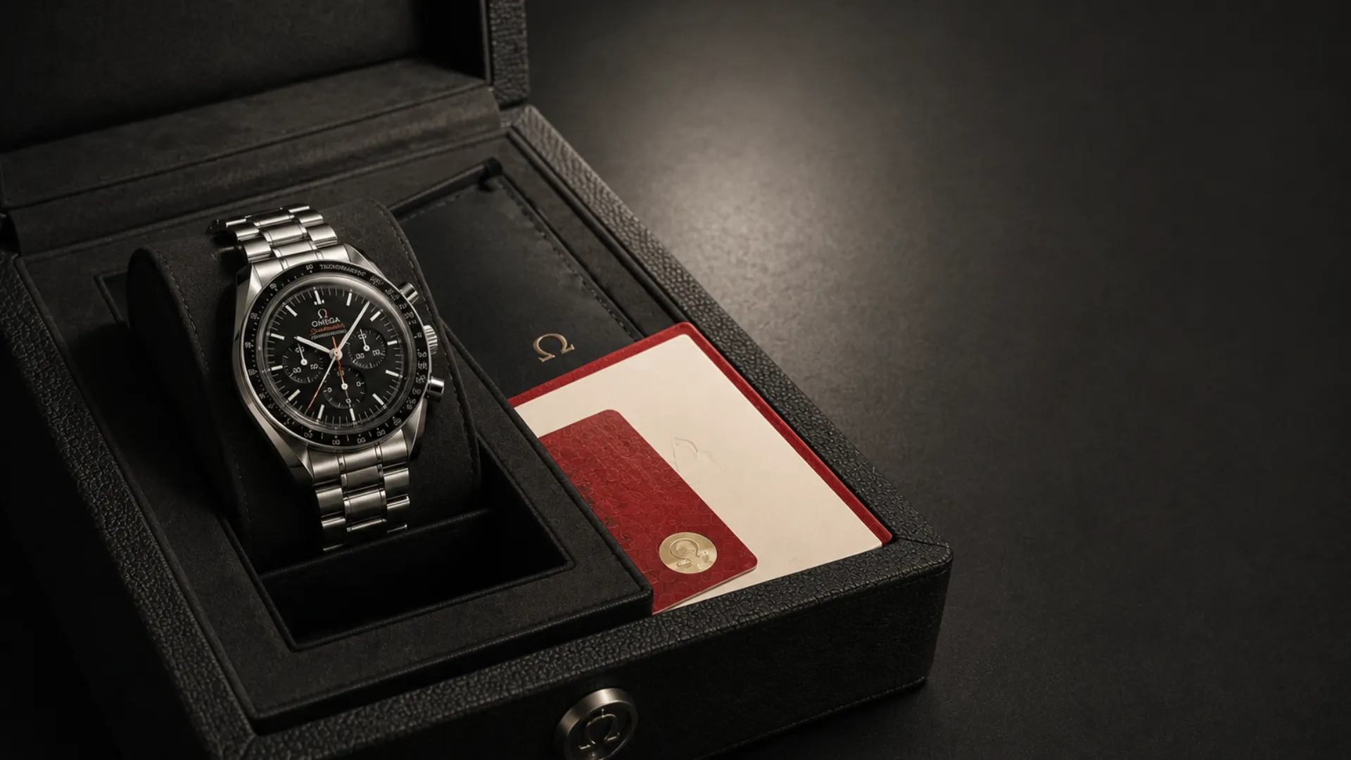 Omega Speedmaster with black dial in an open box alongside its red warranty card and documents on a dark surface