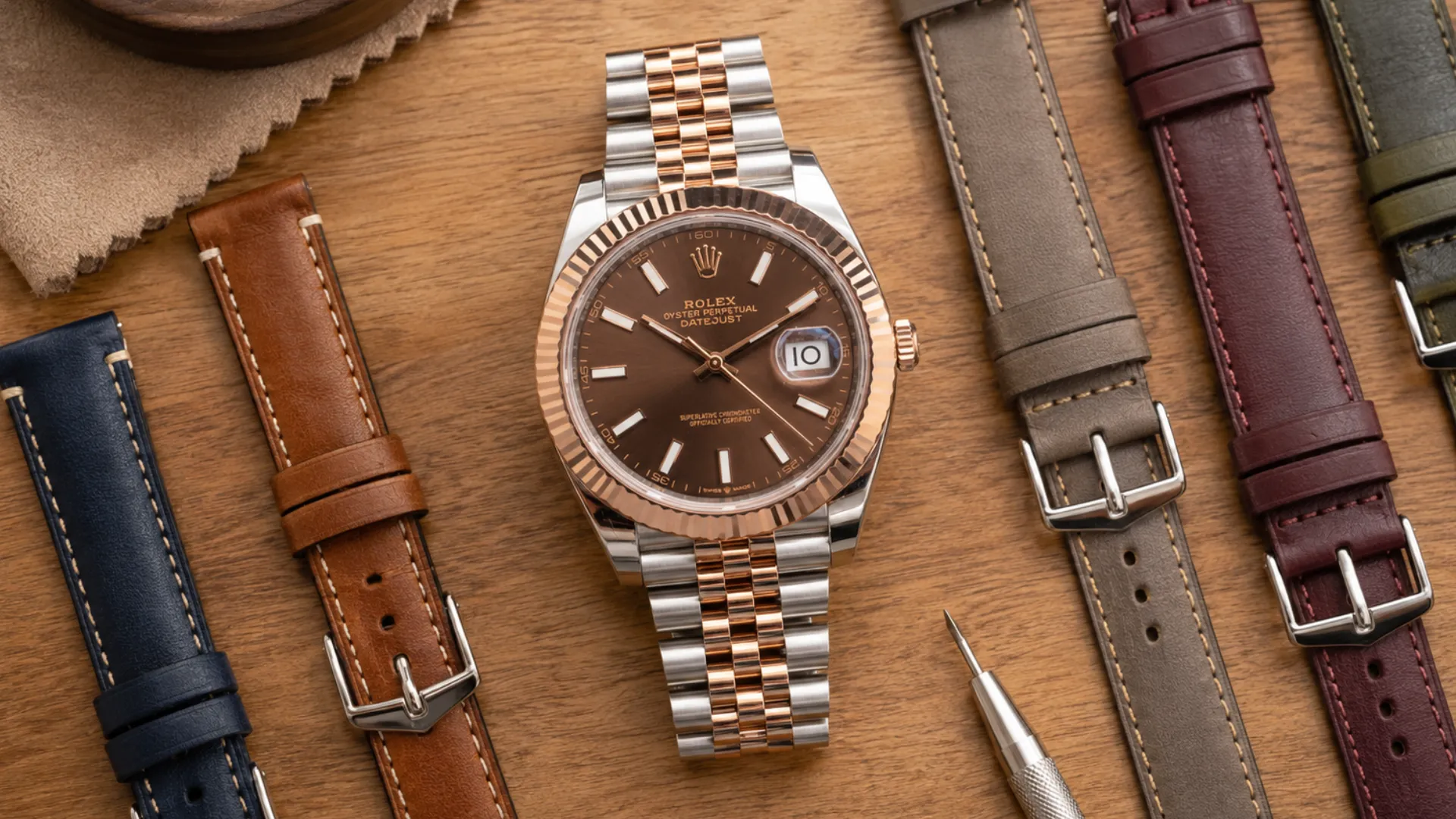 Two-tone rose gold Rolex Datejust with chocolate dial surrounded by navy, tan, taupe, burgundy and green leather straps on a wood surface