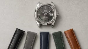 White gold Rolex Datejust with grey Wimbledon dial above a row of black, grey, navy, green, and tan leather straps on a stone surface